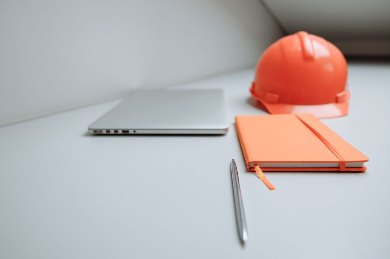 services-04 A minimalist office setup featuring a laptop, orange notebook, and safety hard hat.